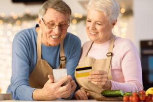 Senior Spouses Shopping Online With Smartphone In Kitchen, Focus On Credit Card And Mobile Phone. Modern Grocery Shopping For Older People Concept. Shallow Depth photo