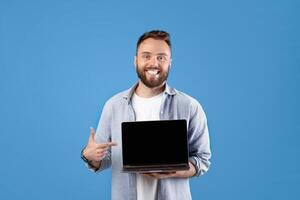 Portrait of happy bearded man holding laptop computer with empty screen over blue studio background, mockup for design. Young guy showing PC with space for your website or advertisement photo