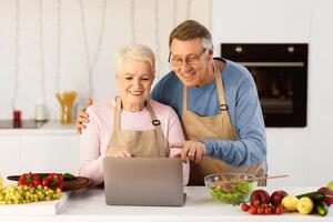 Senior Couple Cooking And Using Laptop Browsing Internet For Online Recipes Preparing Healthy Dinner Together In Modern Kitchen At Home. Aged Spouses Enjoying Food Preparation Using Computer photo
