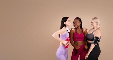 Diverse body positive females in sportswear laughing and posing together, young multi-ethnic ladies standing over beige background in studio, enjoying fitness and healthy lifestyle, copy space photo