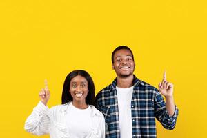 Interesting offer, blogger advice and place for ad. Smiling zoomers african american husband and wife in casual show thumbs up to empty space, isolated on yellow background, studio shot, square photo
