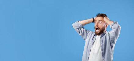 Shocked millennial guy shouting and holding his head in disbelief, looking at empty space on blue studio background, banner design. Overwhelmed bearded man screaming OMG or WOW photo