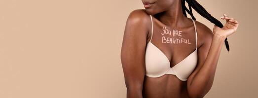 You Are Beautiful. Motivational Inscription Written On Chest Of Unrecognizable Black Woman In Bra, Young African American Female In Underwear Standing Over Beige Background In Studio, Cropped Image photo