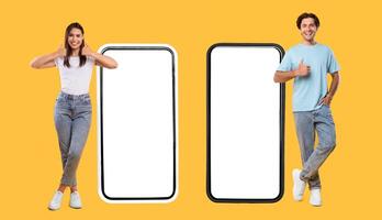 We Like This App. Full body length of happy couple standing next to 2 smart phones with white empty screen for mock up, young lady and guy showing thumb up sign gesture, panorama, banner, free space photo