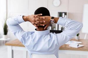 Back view of black businessman in formal shirt sitting at workplace in his office, sitting in chair and leaning back, thinking about something or relaxing during difficult working day, copy space photo