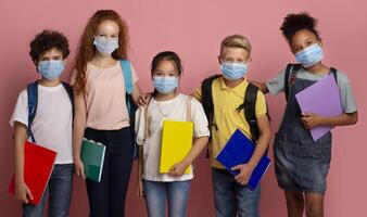 Virus epidemic and schooling. Multiethnic kids in medical masks holding notebooks on pink background photo