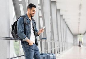 Portrait Of Smiling Arab Male Traveller With Smartphone And Luggage Waiting In Airport, Happy Young Middle Eatern Man Browsing Internet On Mobile Phone While Standing In Terminal, Copy Space photo