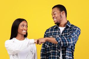 Work as team, agree to something, smile and positive. Funny teens african american students guy and woman in casual give fists bump, isolated on yellow background, empty space, studio shot photo