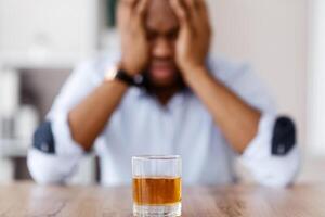 Selective focus on glass of alcohol whiskey over drunk african american office manager or businessman in formal clothes holding his head. Stressed businessman, alcohol addiction concept photo