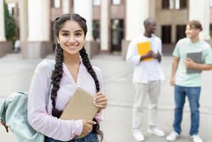 Loans for education concept. Portrait of happy indian female student smiling at camera while posing outdoors in campus with her classmates and university building on background photo