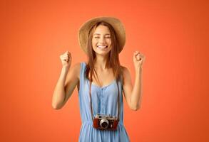 Going vacation. Emotional woman tourist with camera and summer hat raising fists up over yellow studio background, happy to travel. Young excited lady photographer getting ready before travelling photo