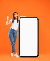 Mobile App Advertisement, Great Offer. Full Body Length Of Excited Lady Pointing Finger Up Leaning On Big Huge White Empty Smartphone Screen Standing On Orange Studio Wall. Cellphone Display Mock Up photo