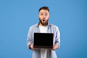 Shocked bearded guy showing laptop with blank screen on blue studio background, mockup for your website. Millennial man demonstrating PC with space for your online advertisement photo