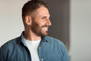 Cheerful middle aged man with stubble smiling while looking aside indoor, portrait shot of charismatic european guy expressing positivity and success. Side view shot photo