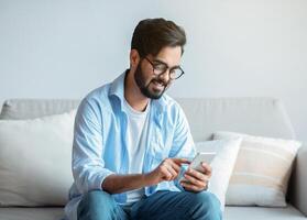 Useful App. Handsome Arab Man In Eyeglasses Using Smatphone While Relaxing On Couch At Home, Eastern Guy Sitting On Sofa In Living Room, Browsing Internet Or Messaging With Friends, Free Space photo
