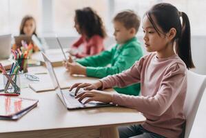 Korean Little Girl Using Laptop Sitting At Desk With Multiracial Classmates In Modern Classroom Indoor. E-Learning Website And Modern Internet Education Service. Selective Focus photo