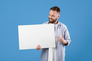 Portrait of positive young guy pointing at blank advertising billboard with mockup for your design. Cheerful bearded man in casual clothes showing empty banner or poster with copy space photo