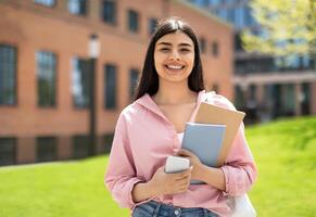 Portrait of happy student girl with cellphone and workbooks walking in college campus and smiling at camera, having break after classes outdoors, copy space photo