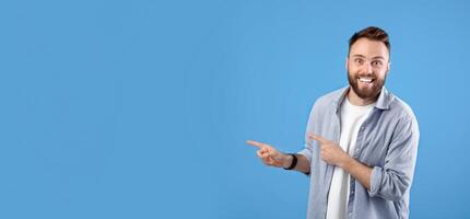 Excited young bearded guy smiling and pointing aside at empty space over blue studio background, panorama. Amazed Caucasian man having unbelievable offer, promoting product photo