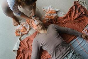 Man lying on the floor while a person ties a cloth around his eyes during a relaxation or meditation practice in a serene indoor setting. photo