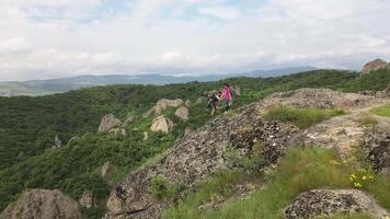 Aerial zoom out two young adult hikers couple reaching hilltop top viewpoint give helping hand through teamwork on steep, rocky slope surrounded by lush green forest and dramatic summer landscape video