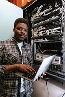 Man working on a laptop in a server room during daylight hours, managing network equipment and data connections photo