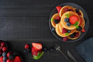 A flatlay image of golden pancakes on a black wooden background, topped with fresh berries create a cozy and indulgent scene. photo