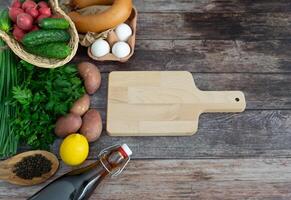 Fresh vegetables and herbs for okroshka styled on a moody dark wood table with a cutting board centerpiece. photo