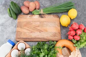 A rustic wooden board anchors the display of fresh okroshka ingredients on a cool gray background. photo