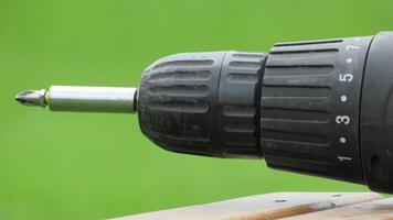 Close-up view of a black electric drill resting on a wooden surface in a green outdoor setting during daylight photo