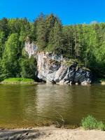 In the center of a flowing river, there is a large rock that is prominently situated, surrounded by a lush forest of trees that line the banks, creating a picturesque scene in nature photo