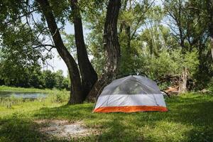 A beautiful and serene camping scene featuring a tent positioned by a tranquil riverbank under the shade of lush trees, creating the perfect spot for relaxation and leisure in natures embrace photo