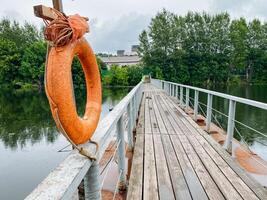 A bright orange lifebuoy sits on an old dock by calm waters, surrounded by lush greenery and colorful foliage, creating a peaceful atmosphere perfect for relaxation and adventure in nature photo