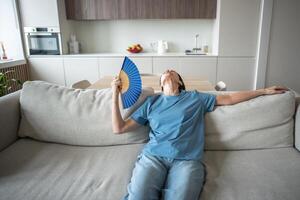 No cooling system, elderly woman struggling without air conditioning, resting on couch using fan photo