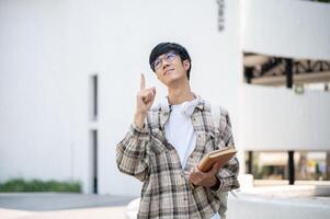 Asian man with glasses holding books and pointing a finger up while standing in front of a building. photo