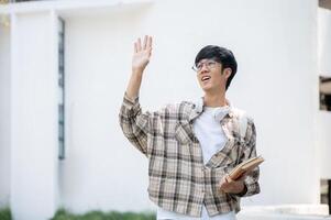 An asian man with glasses is holding books and waving a hand while standing in front of a building. photo