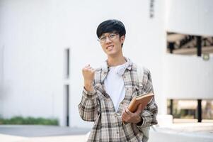An asian man with glasses is holding books and making a fist while standing in front of a building. photo
