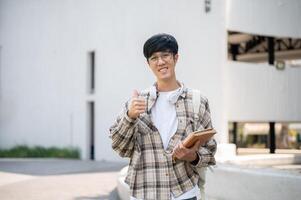 An asian man with glasses is holding books and making thumbs up while standing in front of a building photo