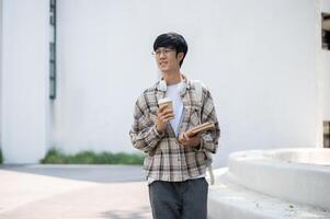 An asian man with glasses is holding coffee with books while standing or walking around the building photo