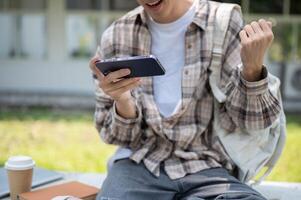 Close up of a man looking at phone and make a fist while sitting with coffee and book on park bench photo