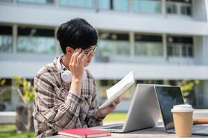 An asian man is touching his glasses while reading a notebook sitting with a laptop at table in park photo