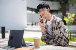 An asian man with glasses is making a call while sitting with laptop at concrete table in park. photo