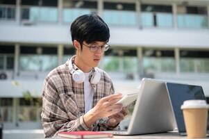 An asian man with glasses is reading a notebook while sitting with a laptop at table in park. photo