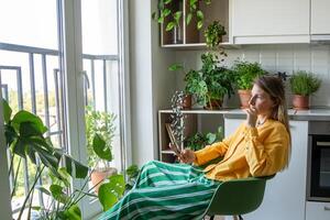 Pensive woman staring out window holding phone in plant filled kitchen. Lost in thoughts, bad news photo