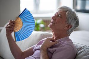 Heat can be deadly for seniors. Sweltering pensioner with fan struggling with summer heat indoors photo