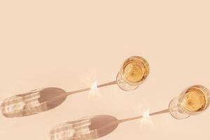 Sunlight still life with two glasses of white wine, beige background with shadow and glare, caustic light effect at sun. Minimal aesthetic trend photo with summer alcohol drink, dark light