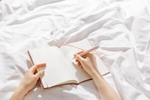 Woman makes plan, day results on blank white page diary journal on bed, writing in notebook or wish diary, holds notepad and pen in hands, Increasing mindfulness, relax and comfortable concept photo