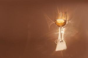 Sunbeam falls on glass of white wine, drink sparkles in sunlight, still life with wineglass of shiny alcohol, beige background with shadow and sun glare, Minimal aesthetic creative photo