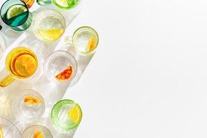 Top view colored Glasses of Drinking natural water with slises of lemon, orange, tangerine, shadow and sunlight reflection on white table, vivid modern flat lay, summer party concept, nonalcoholic photo