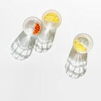 Transparent glass cups of infused water with slices of fruit, clear glasses of detox drink with beautiful shadow at sunlight, minimal aesthetic top view, creative summer drink backdround, above photo
